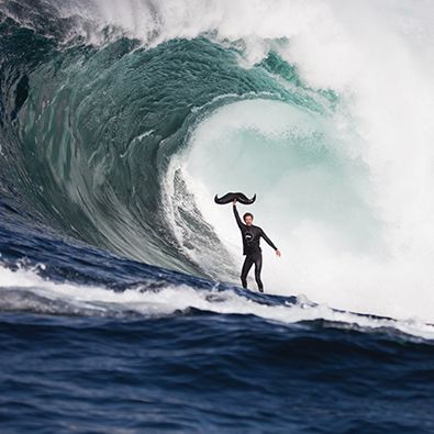 Spectacular photo of a surfer underneath a huge wave, holding a Movember-logo moustache.